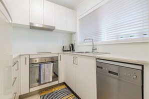 Eine kleine Küche mit weißen Schränken, einem Edelstahlbackofen, einem Geschirrspüler und einem Fenster mit Jalousien.
