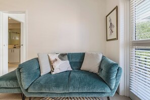 A teal sofa with three cushions in a room with white walls, a framed picture, and a window with blinds. - Botanic Edge Beachside Comfort by St Kildas Icons (Saint Kilda)