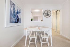 A dining area with a round table and four chairs, a clock on the wall, and a framed abstract painting. - Botanic Edge Beachside Comfort by St Kildas Icons (Saint Kilda)