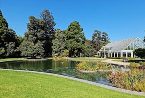 A park with a pond, lush greenery, and a conservatory. - Botanic Edge Beachside Comfort by St Kildas Icons (Saint Kilda)