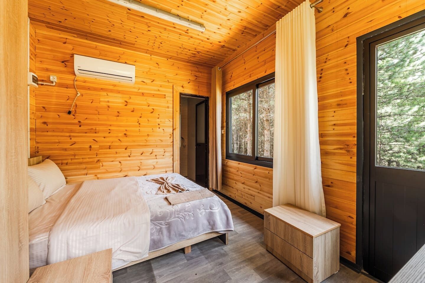 A wooden cabin room with a bed, a small table, a bench, and a window with a view of trees.