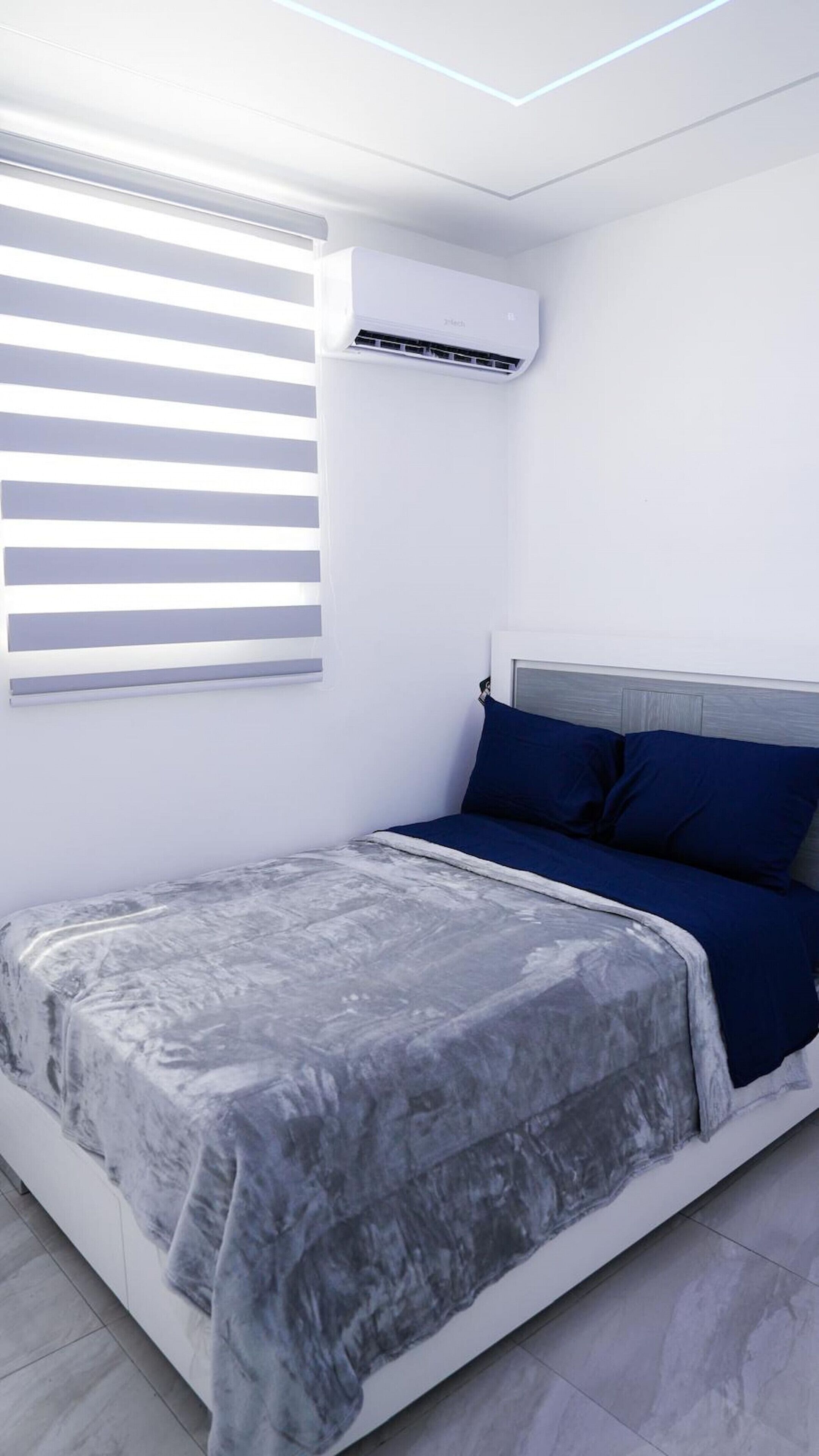 A bedroom with a bed, a window with blinds, an air conditioning unit, and a light fixture.