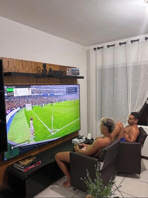 A living room with a flat-screen TV displaying a soccer game, two people watching, a bookshelf, and a potted plant. - A cozy retreat in Búzios. Just a few minutes' walk from the resort's best beach. (Armação dos Búzios)