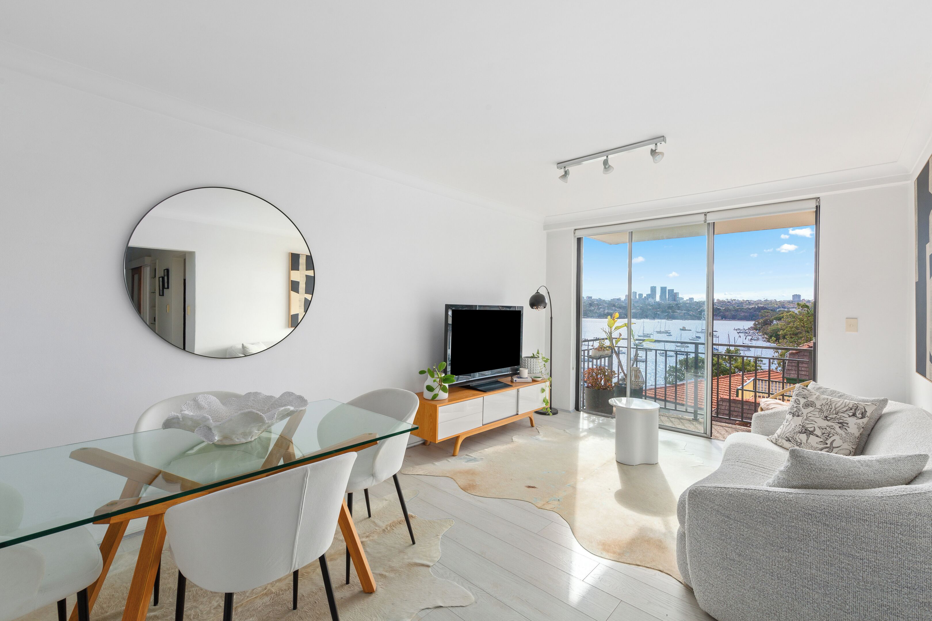 A modern living room with a glass dining table, white chairs, a flat-screen TV, and a sofa.