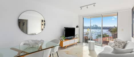 A modern living room with a glass dining table, white chairs, a flat-screen TV, and a sofa.
