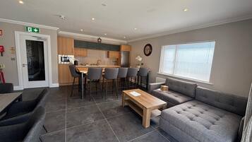 A modern living room with a grey sofa, a wooden coffee table, and a kitchen area with a dining set.