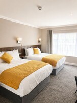 A hotel room with two beds, a headboard, a nightstand, a lamp, a window with curtains, and a carpeted floor.