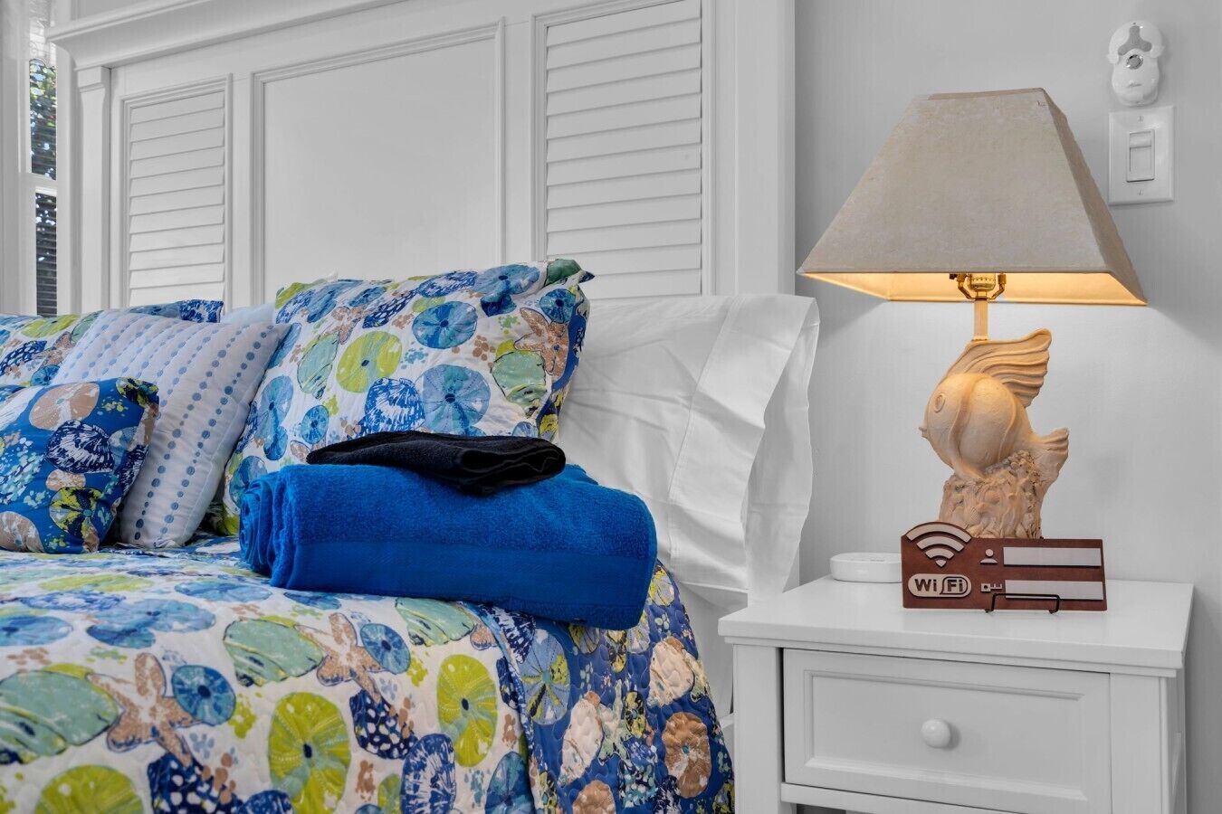 Een bed met bloemenbeddels, een blauwe handdoek en een decoratieve vislamp op een nachtkastje.