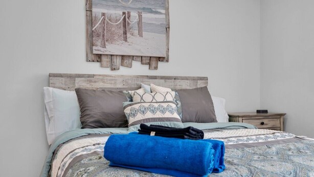 Un lit avec une couvre-lit à motifs, des oreillers et une serviette bleue. Il y a une table de chevet avec une lampe et une petite table. Un tableau représentant un paysage enneigé est accroché au-dessus du lit.