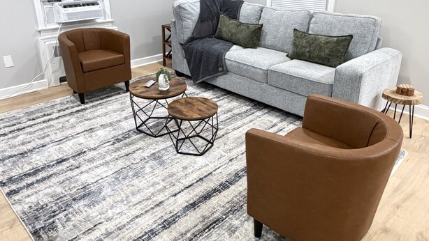 Un salon avec un canapé gris, deux fauteuils marron, une table basse et un tapis à motifs.
