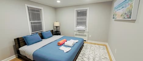 A bedroom with a bed, two bedside tables, a lamp, an air conditioning unit, a patterned rug, and a painting on the wall.