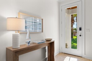 A modern interior with a wooden console table, a lamp, a framed picture, and a door leading to a garden. - Sleeps 10 l 3 Min to Beach l Pool l Game Room l Wireless Internet l Pompano Joes (Miramar Beach)