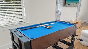 Una taula de futbolí a una habitació amb finestra, una taula petita i una obra d'art penjada a la paret.