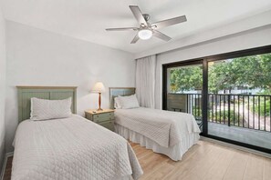 A bedroom with two beds, a ceiling fan, a nightstand with a lamp, and a large window with a view of greenery. - D-226 - Peaceful Views on Quiet End of Resort (Naples)