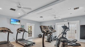 Gimnasio con cintas de correr, elíptica y bicicleta estática, que incluye un televisor, carteles motivacionales y un ventilador de techo.