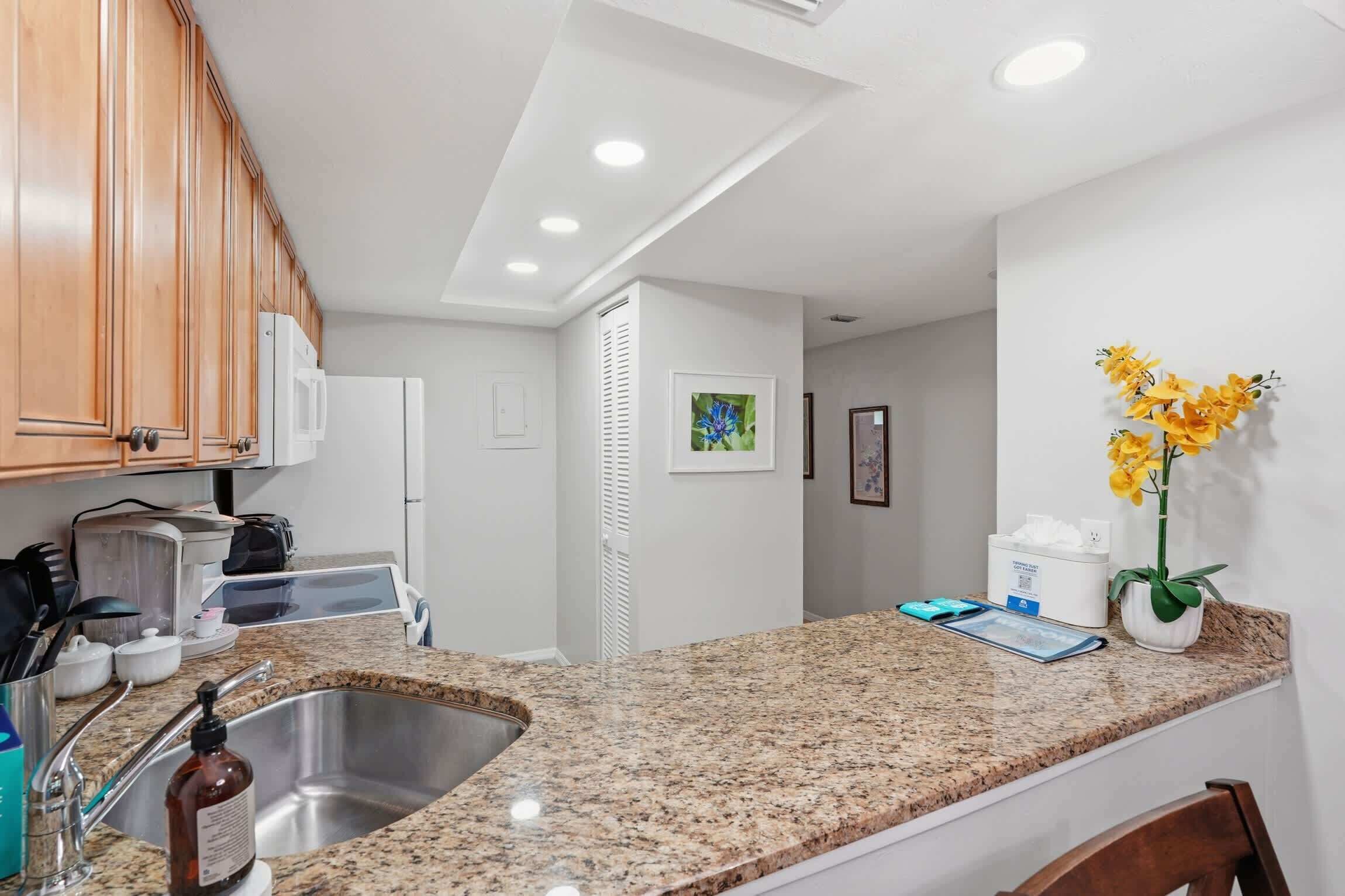 Una cocina con encimeras de granito, pileta de acero inoxidable, muebles de madera y un refrigerador blanco.