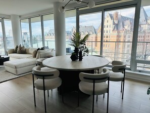 A modern living room with a round dining table, white sofas, and chairs, large windows with a city view. - The Falls - Downtown Victoria, where luxury living meets panoramic views. (Victoria)