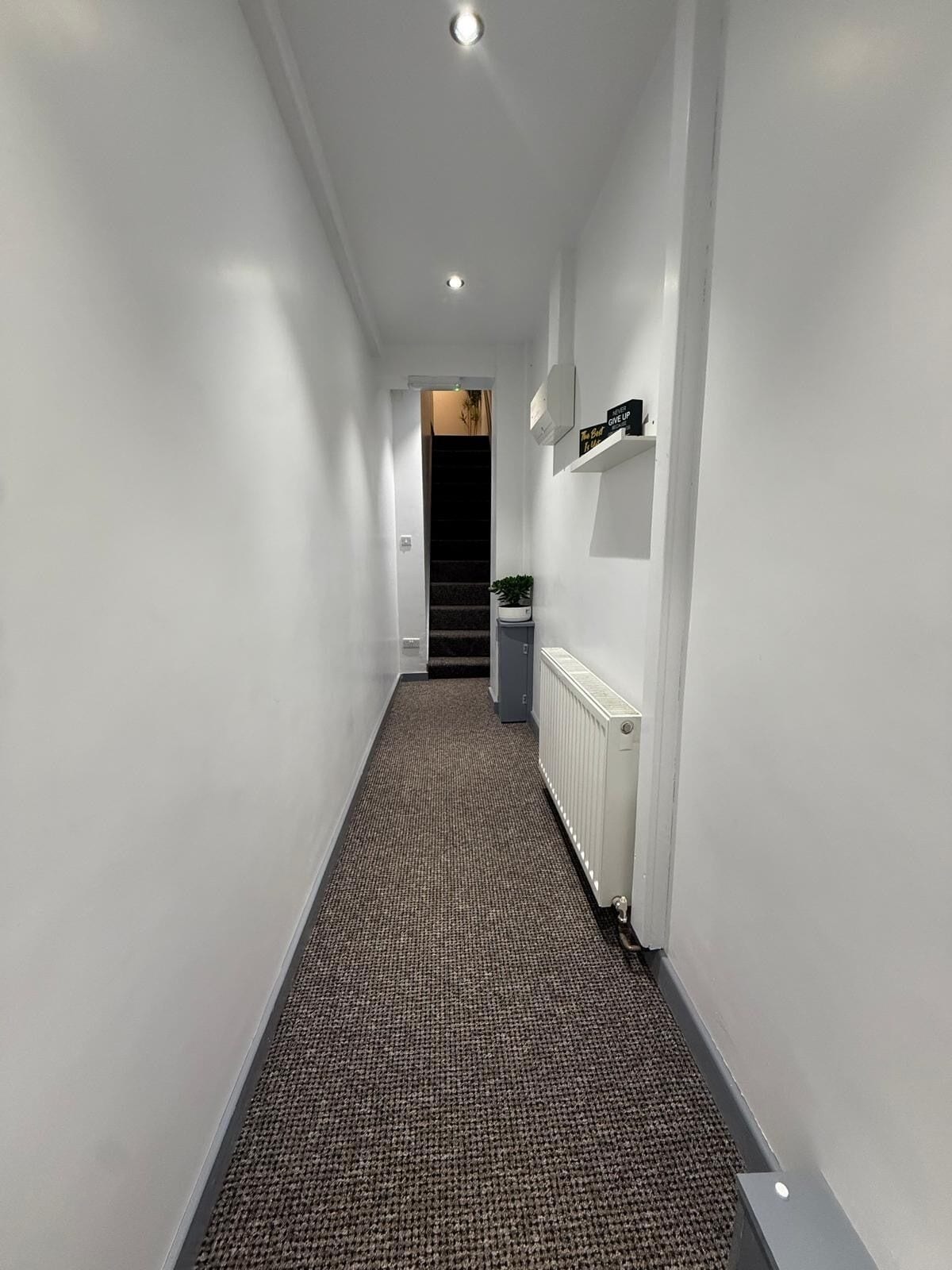 A narrow hallway with a carpeted floor, white walls, and a radiator on the right.