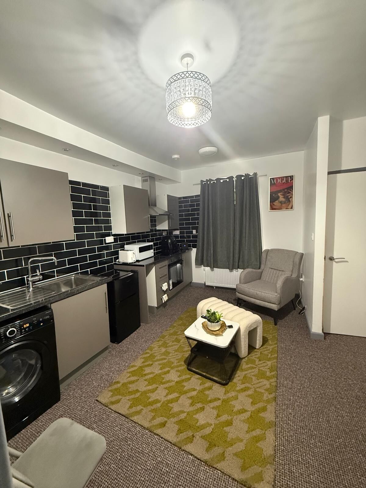A compact living space with a kitchenette, a washing machine, a grey armchair, and a small coffee table.