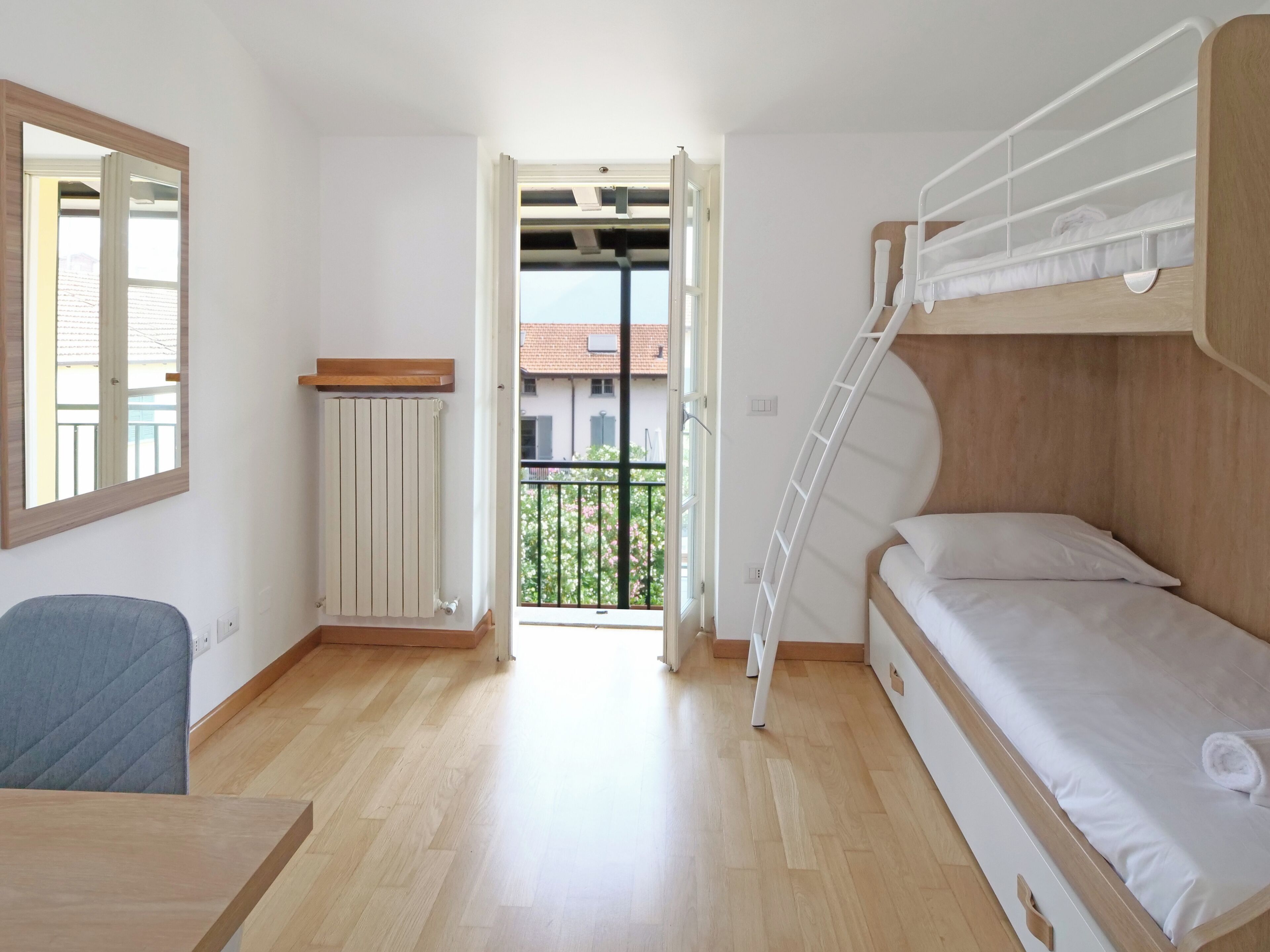 Een kleine kamer met een stapelbed, een bureau, een stoel, een radiator en uitzicht op het buitenbalkon.
