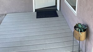 A white door with a small window above it, a black doormat with 'welcome' written on it, and a potted plant on a metal stand to the right.