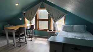 A room with a sloped ceiling, a bed with white bedding, a desk with a chair, and a window with curtains.