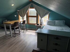 A room with a sloped ceiling, a bed with white bedding, a desk with a chair, and a window with curtains.
