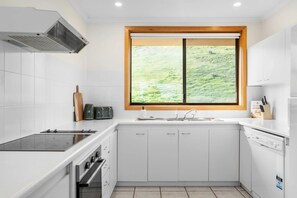 A modern kitchen with white cabinetry, a built-in oven, a stovetop, and a window with a view of greenery. - Carrickalinga House A Symphony of Sea and Serenity (Carrickalinga)