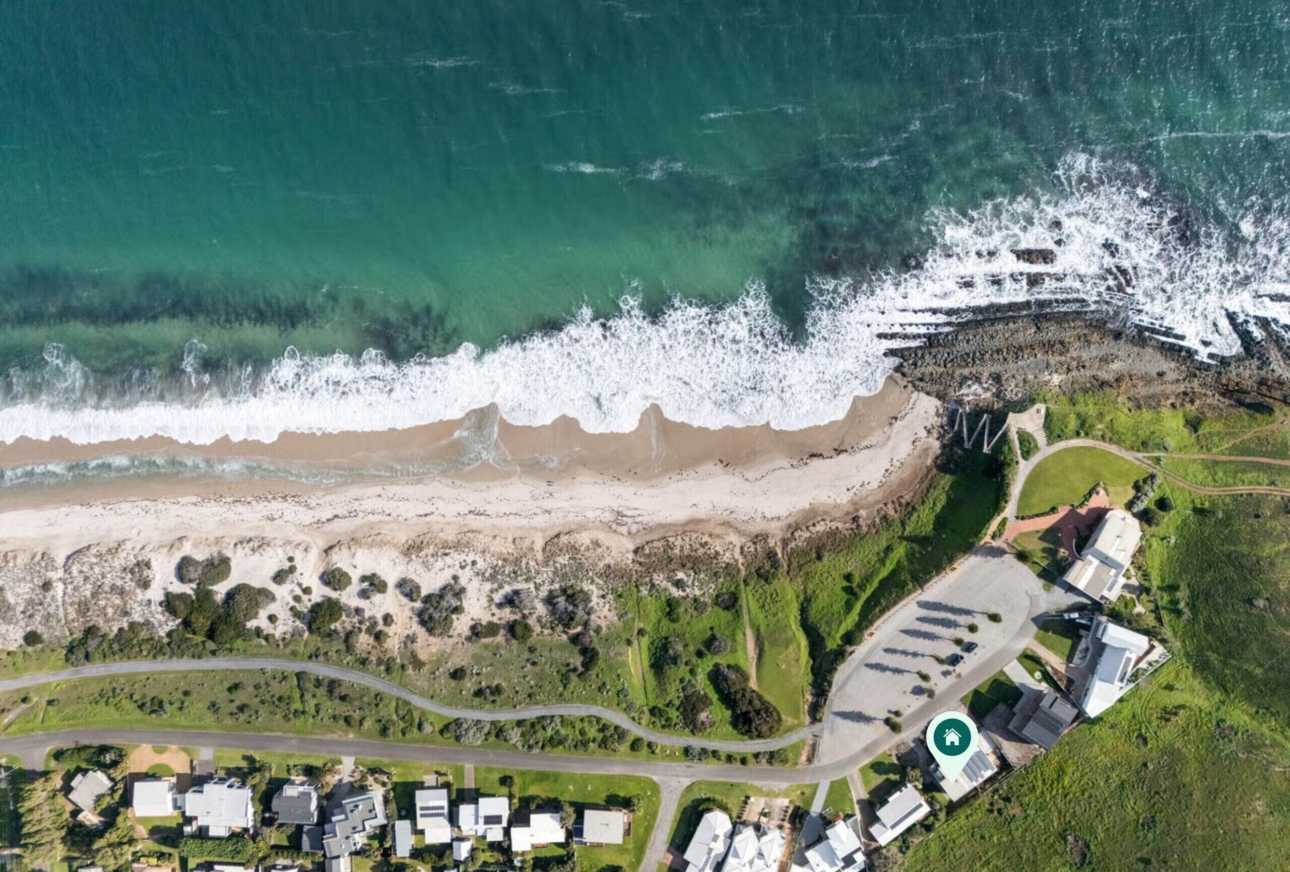 Vue aérienne d’une zone côtière comprenant une plage de sable, une côte rocheuse et une zone résidentielle avec des maisons et un parking.