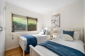 A bedroom with two beds, a window with a view of greenery, a bedside table with a lamp, and a wall-mounted artwork. - Carrickalinga House A Symphony of Sea and Serenity (Carrickalinga)