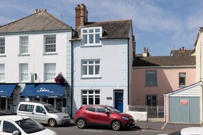Une vue de rue avec un bâtiment blanc doté d’un auvent bleu sur lequel on peut lire l’inscription « John’s », ainsi qu’une enseigne indiquant « Fromages, vins, spécialités locales ». Une voiture rouge est garée devant le bâtiment.