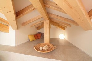 Ein Zimmer mit geneigter Decke, Holzbalken, einem gewebten Korb am Boden und einem gemusterten Kissen.