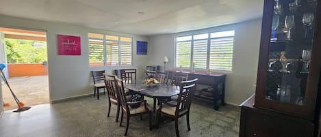A dining area with a round table and chairs, a cabinet with glassware, and two windows with blinds.