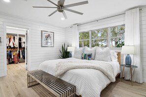 Ein Schlafzimmer mit einem Bett, einem Deckenventilator, einem Fenster mit Vorhängen, einem Nachttisch mit Lampe und einem Kleiderschrank mit Kleidung.