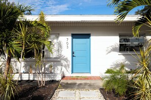 Ein weißes Gebäude mit blauer Tür, Hausnummer 703, umgeben von Palmen und einem kleinen Garten.