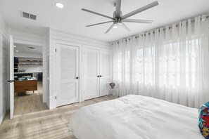 Ein Schlafzimmer mit einem Bett, einem Deckenventilator, weißen Vorhängen und Blick auf einen Küchenbereich.