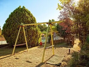 Ein Spielplatz mit Schaukeln und Klettergerüsten, umgeben von Bäumen und einem Zaun.