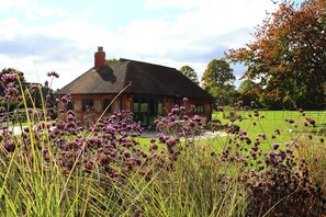 A brick house with a sloped roof and a chimney, surrounded by a grassy field and purple flowering plants. - Beautiful Cottage with Tennis Court and Games Barn (Hambleden)