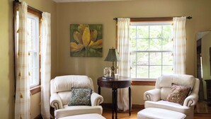 A room with two white armchairs, a small table with a lamp, and a painting on the wall.