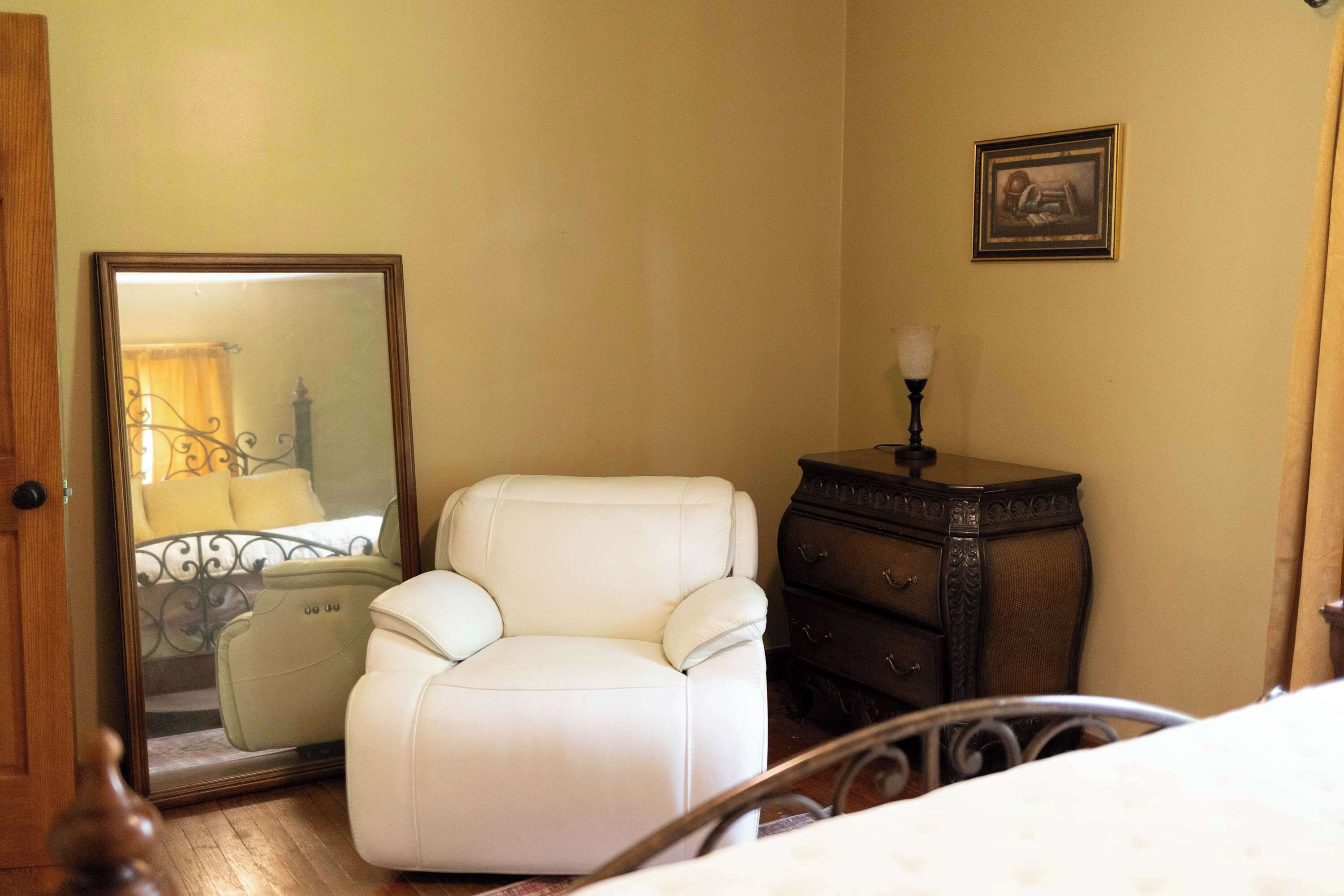 A bedroom with a bed, a white armchair, a dresser, a mirror, and a framed picture on the wall.