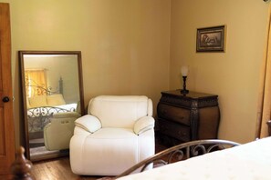 A bedroom with a bed, a white armchair, a dresser, a mirror, and a framed picture on the wall.