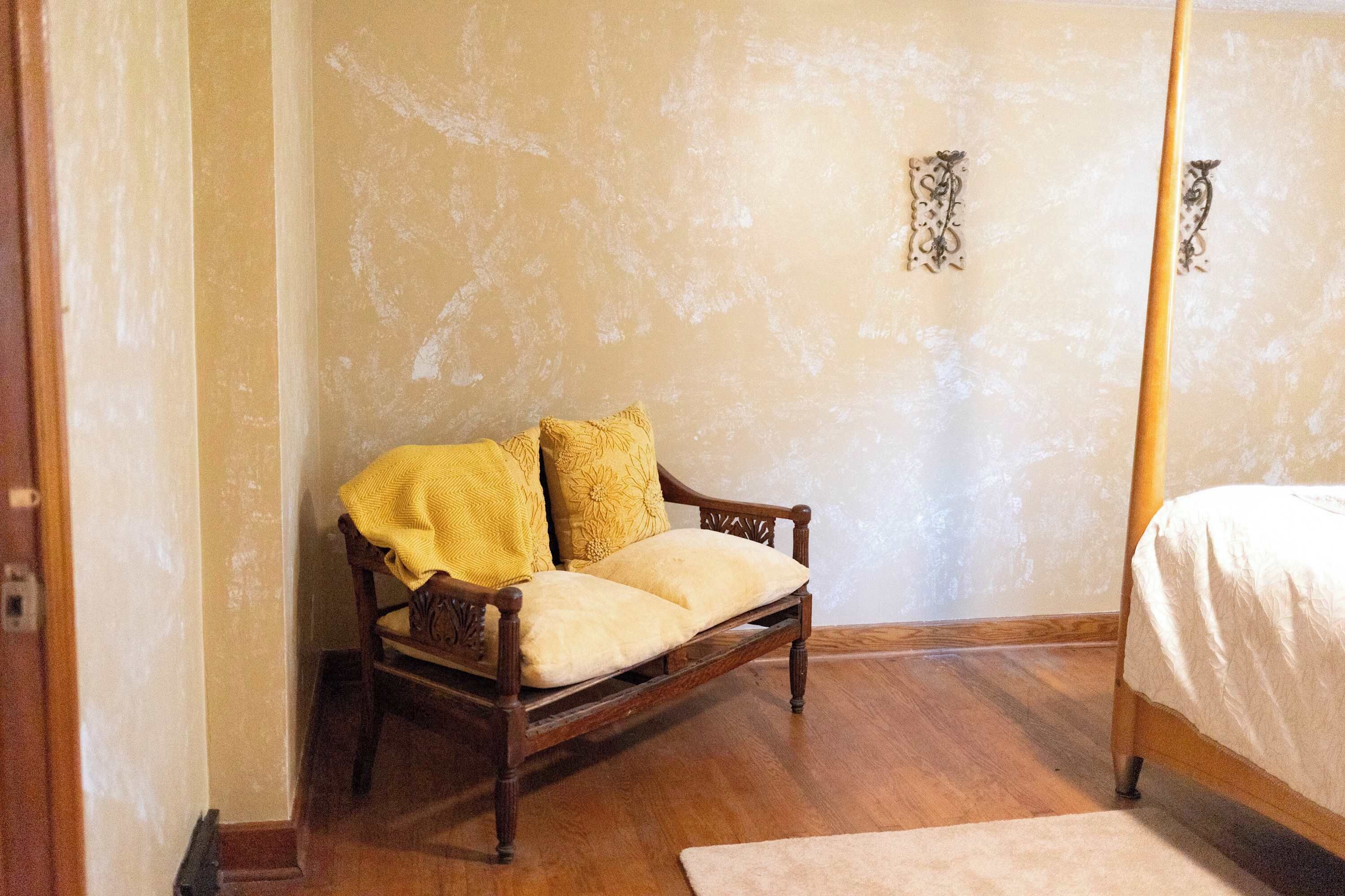 A bedroom with a wooden four-poster bed, a wooden chair with cushions, and a plain wall with a small decorative item.