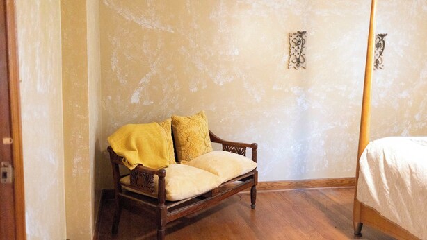 A bedroom with a wooden four-poster bed, a wooden chair with cushions, and a plain wall with a small decorative item.