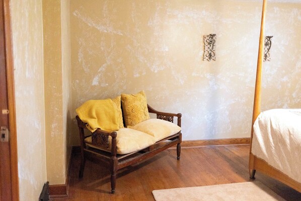 Un dormitorio con una cama con dosel de madera, una silla de madera con cojines y una pared lisa con un pequeño adorno.