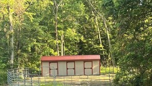 A fenced area with a red building, possibly a barn or stable, surrounded by trees.