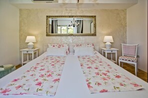 Un lit double avec une literie à motifs floraux, deux tables de chevet avec des lampes, une chaise et un grand miroir au mur.