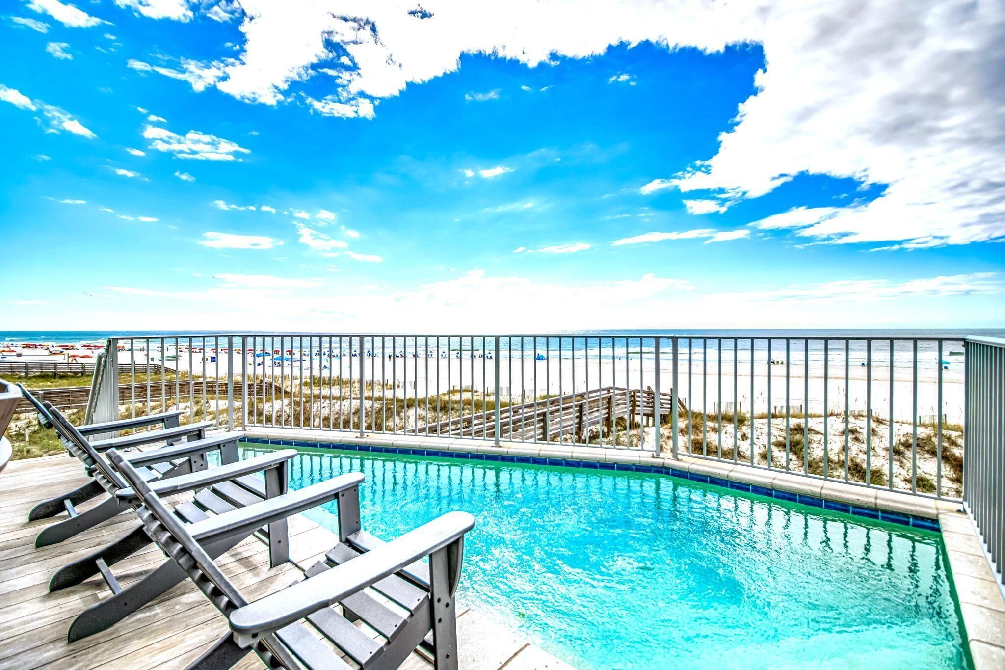 A swimming pool with a wooden deck and lounge chairs, surrounded by a metal fence, overlooking a beach with umbrellas.