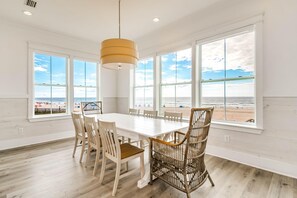 A spacious dining area with a long table and chairs, large windows with a beach view, and a hanging light fixture. - Beachfront Bliss Beach House (Orange Beach)