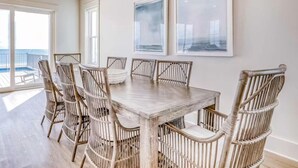 A dining area with a wooden table and wicker chairs, large windows, and framed artwork on the wall. - Beach Vista Two Beach House (Orange Beach)