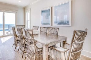 A dining area with a wooden table and wicker chairs, large windows, and framed artwork on the wall. - Beach Vista Two Beach House (Orange Beach)